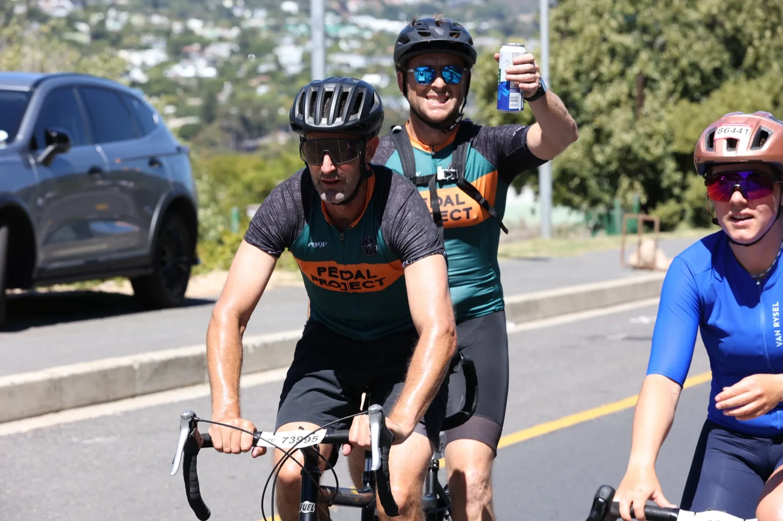 Two Pedal for Good riders on the road during the Cape Town Cycle Tour, one raising an arm to the camera.